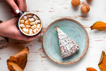 Top view of female hands holding marshmallows on shabby table with piece of cake, walnuts and scattered leaves