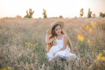 Naklejka na ściany i meble Cute little girl with blond long hair in a summer field at sunset with a white dress with a straw hat