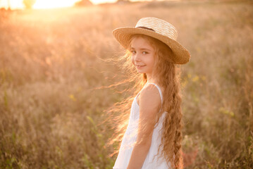 Cute little girl with blond long hair in a summer field at sunset with a white dress with a straw hat