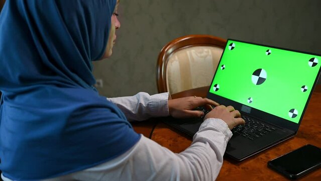 Rear view of a confident multitasking Arab Muslim woman in hijab, sitting at wooden desk and telecommuting, typing text on laptop with green chroma key mockup screen for insert promotional clip, video