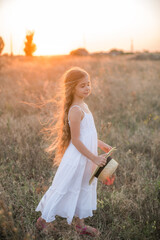 Cute little girl with blond long hair in a summer field at sunset with a white dress with a straw hat