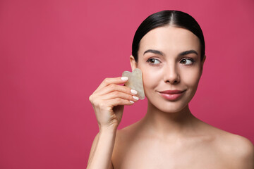 Beautiful young woman doing facial massage with gua sha tool on pink background. Space for text