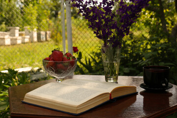 Open book, cup of tea, strawberries and beautiful wildflowers on table in garden