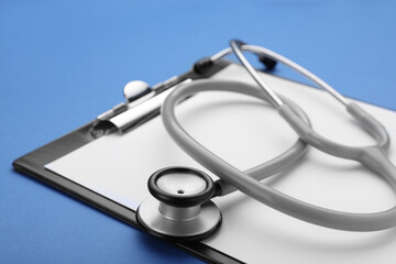 Clipboard with stethoscope on blue background, closeup