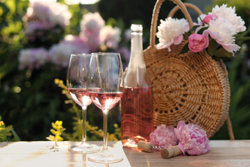 Bottle and glasses of rose wine near beautiful peonies on wooden table in garden
