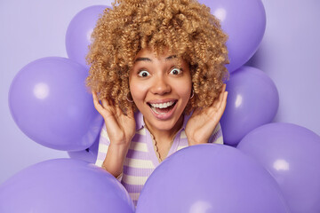 Horizontal shot of surprised cheerful woman with curly blonde hair looks amazed at camera stares impressed smiles broadly cannot believe own eyes poses around inflated balloons against purple wall