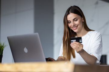 Portrait of a young woman holding credit card while sitting with laptop computer at the table at home