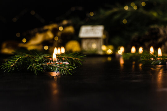 Under the Christmas tree are gifts and small candles are burning. A wooden house and the lights of a Christmas garland are visible against a blurred background. Christmas home decoration concept.