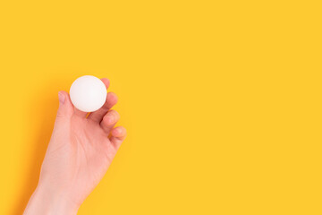 Female hand hold white ball for ping pong on a yellow background. Minimalistic composition with place for text. - obrazy, fototapety, plakaty