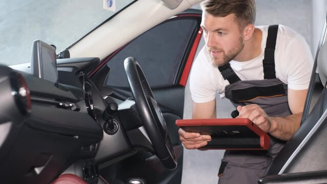 A young car mechanic of American appearance checks the technical condition of the car using a tablet.