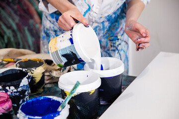 Cropped photo of hands of woman in robe covered with stains pouring white acrylic paint from bucket into other bucket.
