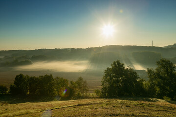 A view over a valley in the rolling hill landscape outside Maastricht, where the valley is covered with layers of fog and creating a nice setting with the sun beams through the mist over the fields