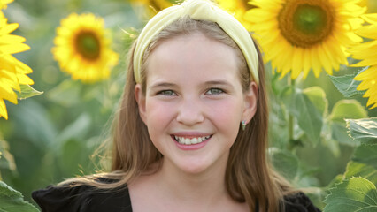 Teenager girl hides behind bright sunflowers among field showing toothy smile. Schoolgirl wearing yellow headband enjoys walking on field