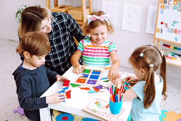 Little kids and educator studying, folding cubes and paper geometric figures on desk sitting on floor in playroom. Interesting lesson for kindergartners developing logic and intelligence - obrazy, fototapety, plakaty