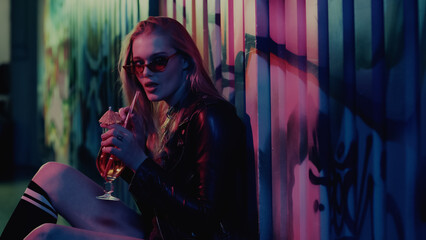 Stylish woman in sunglasses holding cocktail near wall with graffiti in nightclub.