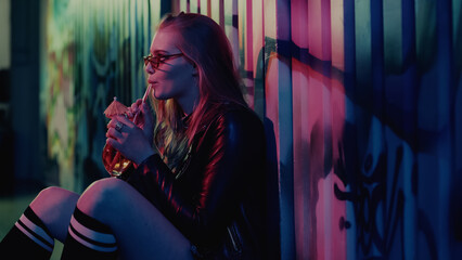 Young woman in sunglasses drinking cocktail near wall with graffiti during party.
