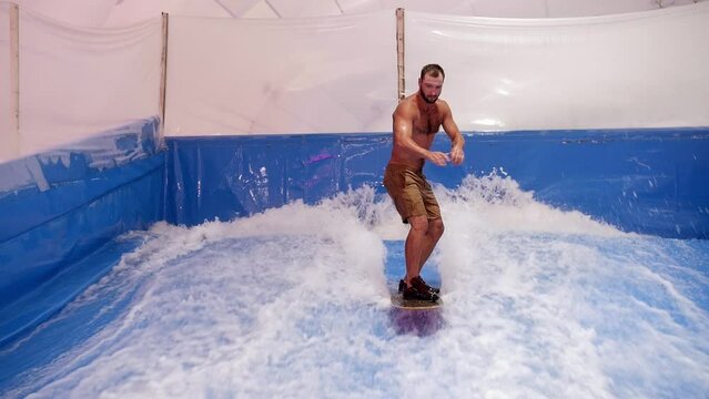 Attractive Man riding surf board on wave simulator indoors. Young surfer during training on generated waves. Water sport activity. Surfer guy enjoy surfing on indoor surf imitator. Surfing concept 