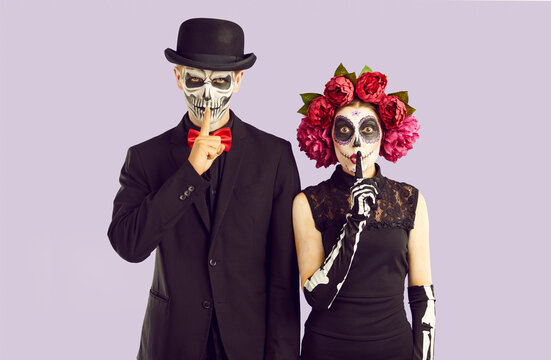 Studio portrait of couple dressed up for Halloween or Day of the Dead. Funny male and female model in spooky costumes and with skull makeup put fingers to lips asking you to be silent and keep secret