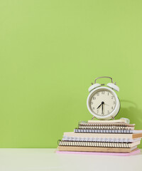 Round alarm clock stands on a stack of notebooks, green background.