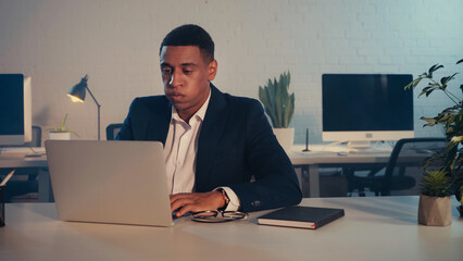 Exhausted african american manager using laptop near eyeglasses and notebook in office at night.