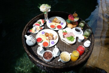 Set of floating breakfast tray in swimming pool with fried egg omelette, sausage, ham, bread, fruit, juices, and other. Breakfast in tropical swimming pool, breakfast set in float tray in resort hotel