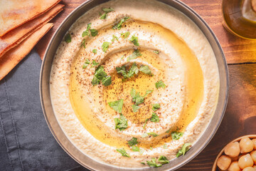 Arabic cuisine; Traditional creamy hummus dip in dark bowl. Topped with olive oil, paprika and parsley. Top view with close up.