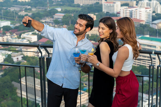 International group of male and female friends holding a smartphone selfie with a girl friend holding a glass of cocktails, party and restaurant on the roof of the building.