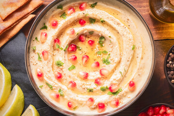 Arabic cuisine; Traditional creamy hummus dip in dark bowl. Topped with olive oil, fresh pomegranate seeds and parsley. Top view with close up.
