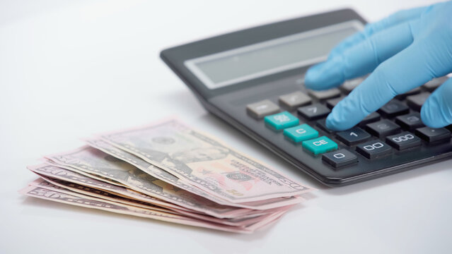 cropped view of person in latex glove using calculator near dollars on white background.