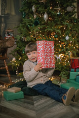 Obraz na płótnie Canvas Pretty little boy sitting in a cozy atmosphere near the Christmas tree. Opening Christmas presents.