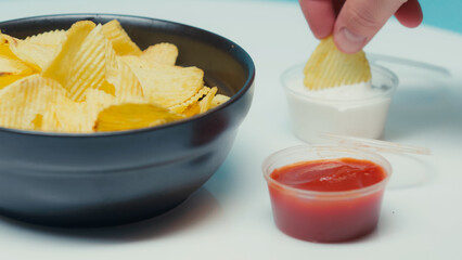 ridged potato chips in bowl near containers with ketchup and mayonnaise on blue.