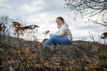 In the spring afternoon, a girl with a scythe sits on the edge of a cliff in blue jeans and a  shirt, side view.