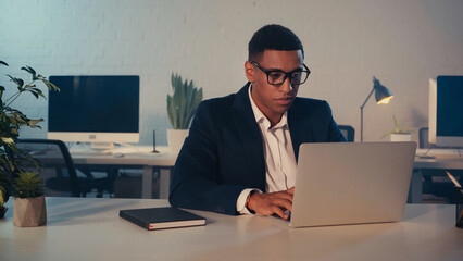 African american manager in eyeglasses using laptop near notebook in office at night.