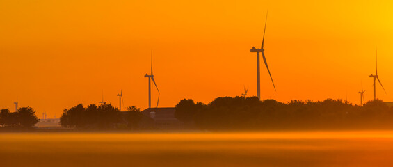 Naklejka na ściany i meble Wind turbines in a foggy agricultural field in sunlight at sunrise in autumn, Almere, Flevoland, Netherlands, September, 2022