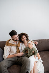 happy parents looking at infant girl in headband while sitting on sofa in living room.