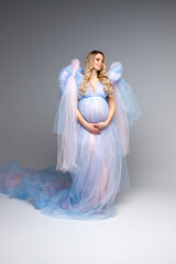 Fototapeta na wymiar A pregnant belly. Pregnant girl with blonde hair on a gray background. Dresses for pregnant women. Maternity clothes. Portrait of a woman in dress. Vertical photo. Angel in blue dress.