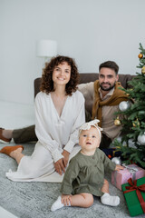 cheerful family with infant baby girl sitting near christmas tree with presents.