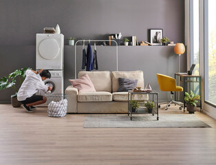 Young man is living at minimalist home style, working laptop, laundry, washing machine, looking phone at the room, grey wall background and decorative interior.