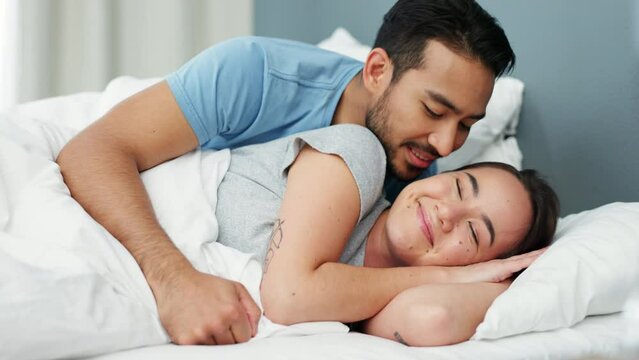 Love, morning and couple waking up in a bed together in their comfortable bedroom at home. Happy, young and calm man giving his wife a kiss to wake up from her sleep in a hotel room on holiday.