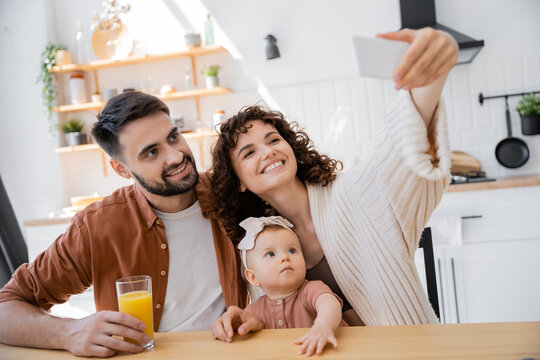 curly woman taking selfie with happy husband and infant daughter at home.