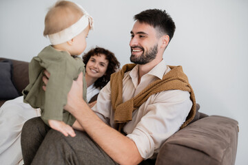 cheerful father holding infant daughter near happy blurred wife in living room.