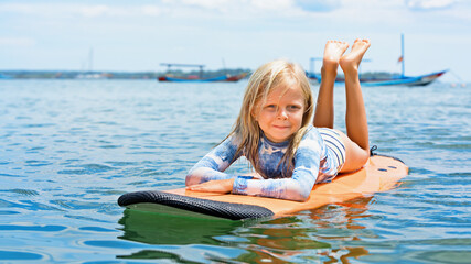 Happy baby girl - young surfer learn to ride on surfboard with fun on sea waves. Active family lifestyle, kids outdoor water sport lessons, swimming activity in surf camp. Summer vacation with child.