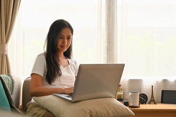 Fototapeta premium Domestic life concept at home with Asian female using laptop while sitting on the sofa in the living room.