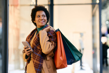 Happy black woman with shopping bags using mobile phone while standing in front of store.