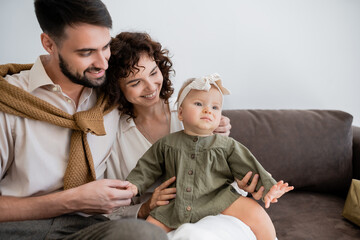 cheerful parents looking at infant girl in headband while sitting on sofa in living room.