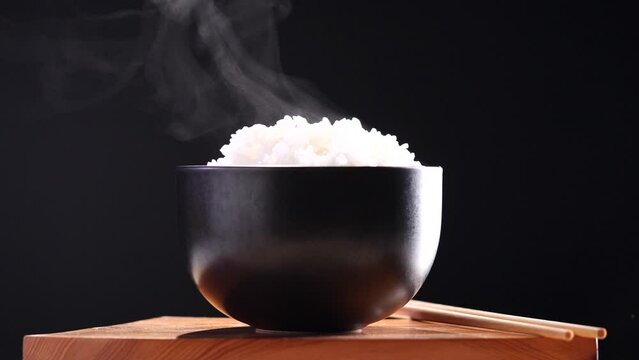 Japanese rice, Cook rice. Close up natural steaming cooked Japanese white rice in black bowl with chopstick on black background, soft focus. Healthy Food Concept.