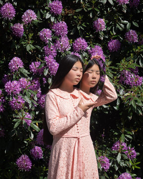 asian woman in pink dress holding mirror reflecting herself near purple rhododendron flowers