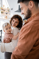 happy mother holding infant baby girl near smiling and blurred husband at home.
