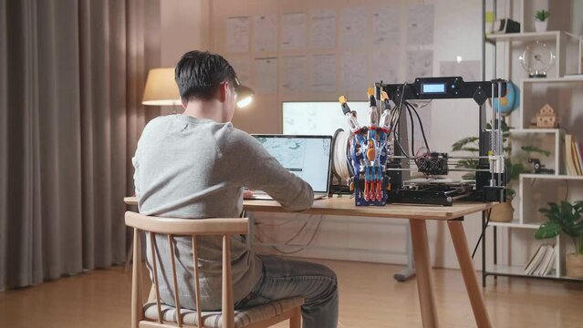 Back View Of A Male With 3D Printing Designing A Cyborg Hand On A Laptop At Home
