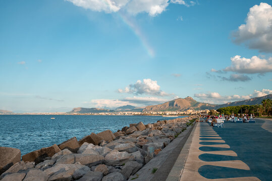 Distant view of tourists relaxing on promenade. Mountains and old town at coastline in background. Rocks arranged at seafront with blue sky in background on sunny day.